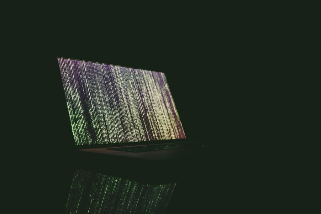 A laptop displaying green and purple code in a dark room, reflecting on a surface.